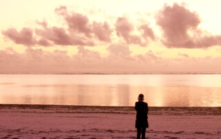 Eine Person steht am Meer und blickt auf den warm beleuchteten Horizont.